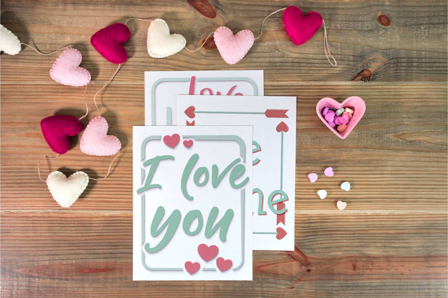 A Valentine's Planner from Organized 31 Shop lies on a wooden surface, surrounded by a garland of fabric hearts. Nearby, a small heart-shaped dish overflows with candy and scattered candy hearts, creating the perfect setting for charming wall art.