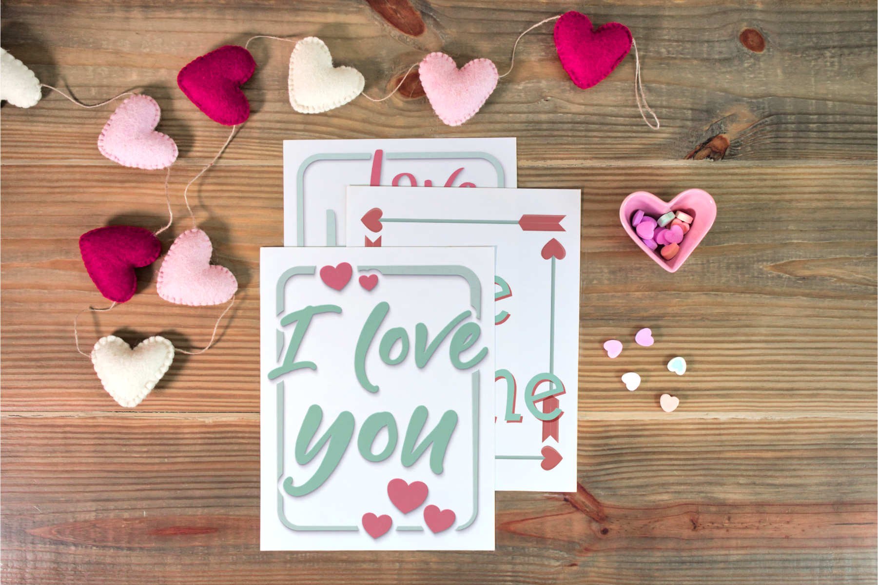 A Valentine's Planner from Organized 31 Shop lies on a wooden surface, surrounded by a garland of fabric hearts. Nearby, a small heart-shaped dish overflows with candy and scattered candy hearts, creating the perfect setting for charming wall art.